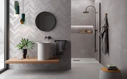 Salle de bain moderne avec murs en **grès cérame** à chevrons et texturés. Lavabo suspendu, douche à l'italienne, miroir rond et touches végétales.