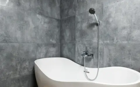 Baignoire blanche aux formes douces, adossée à un mur de carrelage gris en pose en quinconce. Douchette chromée au-dessus.