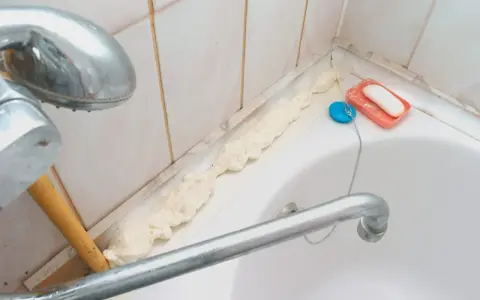 Mousse expansive appliquée pour nettoyer joint plastique porte douche. Savon et bouchon de bain à proximité.