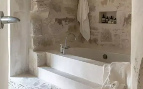 Une **salle de bain méditerranéenne** avec baignoire en pierre brute, robinetterie moderne et serviette blanche suspendue. Ambiance zen.