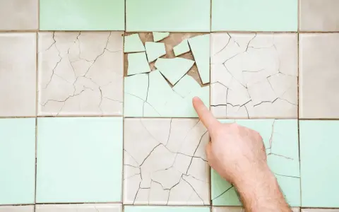 Un doigt pointe une **fissure carrelage** sur un mur de carreaux anciens, certains brisés, d'autres craquelés.