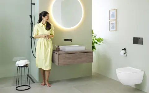 Une femme en robe jaune admire une salle de bain moderne. Le mur vert pâle est une tendance couleur mur toilette, complété par un meuble vasque en bois et un WC suspendu.