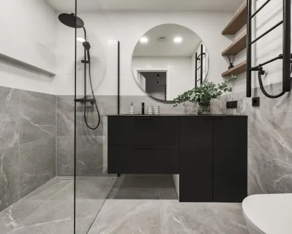 Salle de bain moderne avec douche à l'italienne, meuble suspendu noir, miroir rond et carrelage effet marbre. La construction de cette salle de bain est soignée.