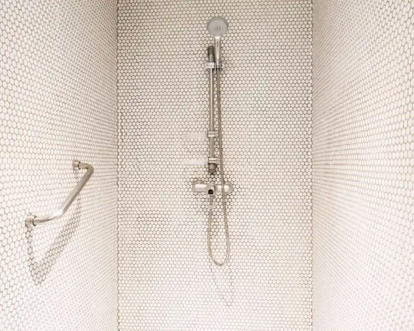 Douche moderne avec carrelage blanc en mosaïque. Une barre d'appui chromée et un pommeau de douche argenté se détachent sur le fond. Le receveur noir est discret.