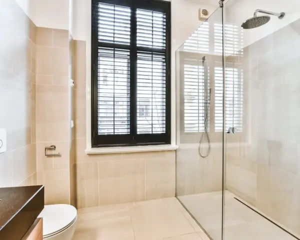 Salle de bain moderne avec douche à l'italienne, carrelage beige, fenêtre à persiennes noires et WC suspendu.