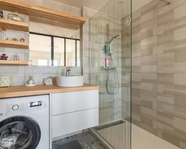 Coin buanderie dans salle de bain moderne avec lave-linge, lavabo, douche vitrée et étagères décoratives.