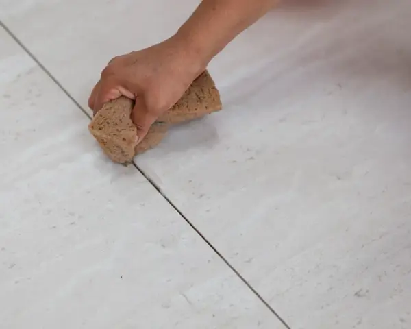 Main tenant, quelle couleur de joint pour carrelage beige ? Une main utilise une éponge pour nettoyer les joints entre des carreaux beiges clairs.