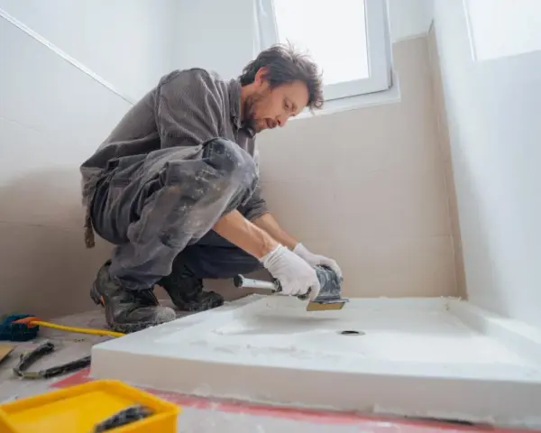 Un ouvrier prépare le sol pour installer un receveur de douche. Il utilise une ponceuse pour assurer une surface plane, essentielle pour le collage.
