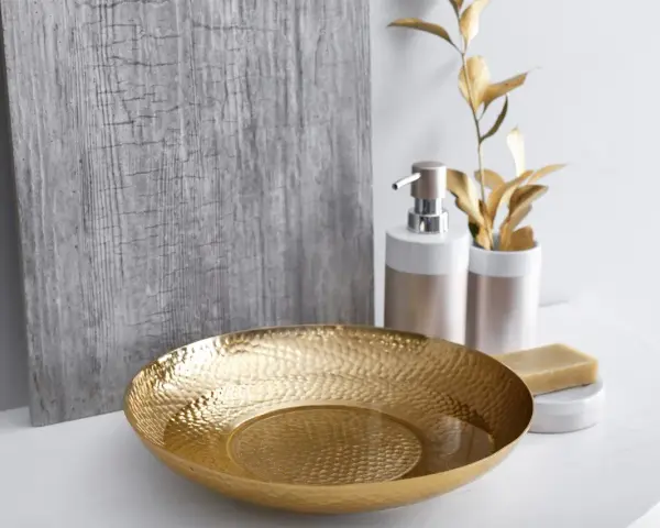 Un bol martelé doré, un distributeur de savon et une barre de savon sur un comptoir blanc, avec une plante dorée et un panneau texturé gris en arrière-plan. Une touche de luxe pour votre douche dorée.