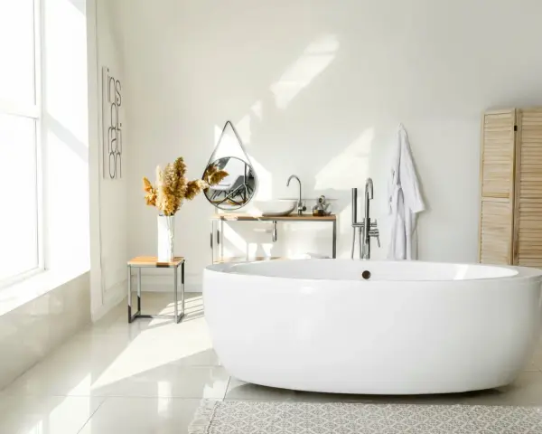Salle de bain lumineuse avec baignoire îlot blanche, lavabo, miroir, porte-serviettes et paravent en bois. La largeur d'une baignoire est parfaite pour ce coin détente.