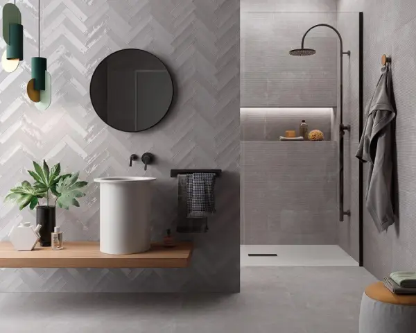 Salle de bain moderne avec murs en **grès cérame** à chevrons et texturés. Lavabo suspendu, douche à l'italienne, miroir rond et touches végétales.