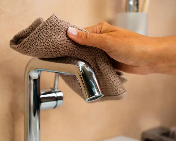 Main d'une femme nettoyant un mitigeur de douche avec un tissu marron.