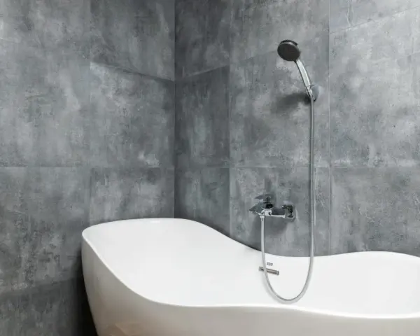 Baignoire blanche aux formes douces, adossée à un mur de carrelage gris en pose en quinconce. Douchette chromée au-dessus.