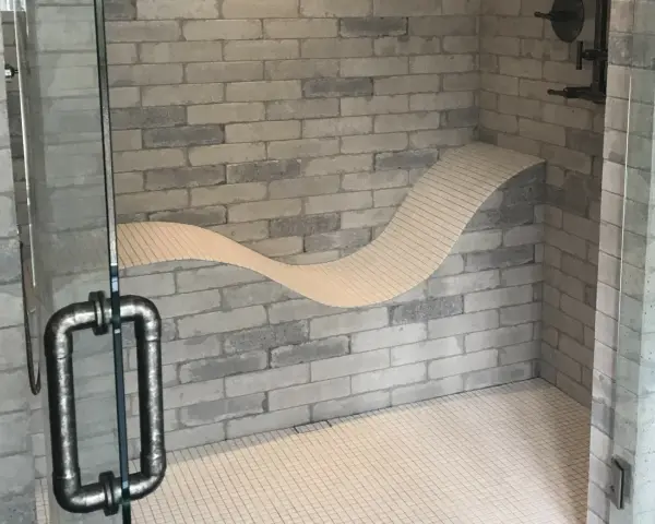 Douche moderne avec un banc ondulé intégré dans le mur de briques. La poignée de la porte en verre est en métal vieilli.