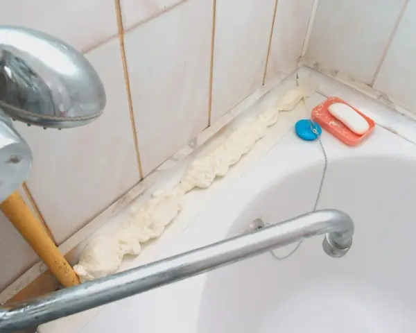 Mousse expansive appliquée pour nettoyer joint plastique porte douche. Savon et bouchon de bain à proximité.