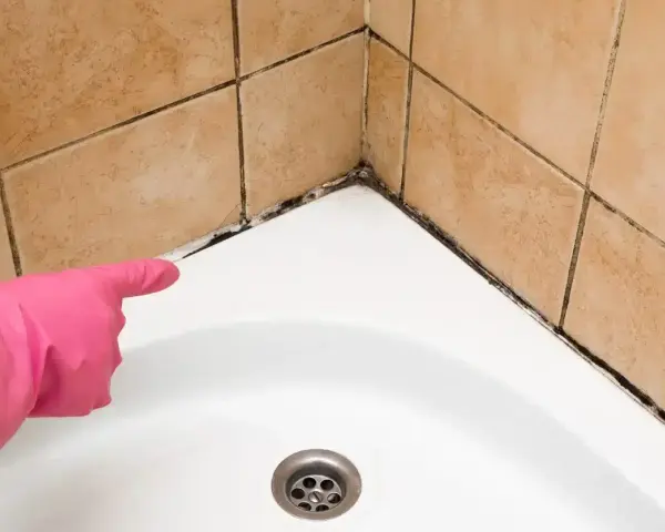 Main d'une personne portant un gant rose pointe vers le joint de douche moisi, indiquant qu'il faut refaire le joint de douche.