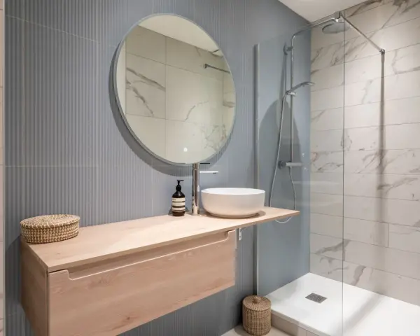 Aménagement petite salle de bain : meuble suspendu en bois clair, vasque ronde, miroir rond, douche à l'italienne.
