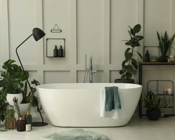 Une salle de bain baignée de lumière, avec une baignoire blanche, de nombreuses plantes vertes qui prospèrent dans l'humidité, et un miroir ovale.