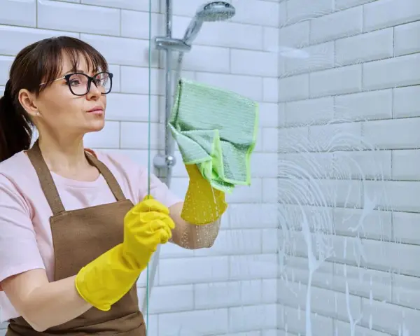Femme avec des gants jaunes et une lingette verte en train de nettoyer le calcaire de la douche.