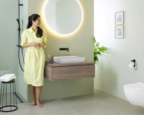Une femme en robe jaune admire une salle de bain moderne. Le mur vert pâle est une tendance couleur mur toilette, complété par un meuble vasque en bois et un WC suspendu.