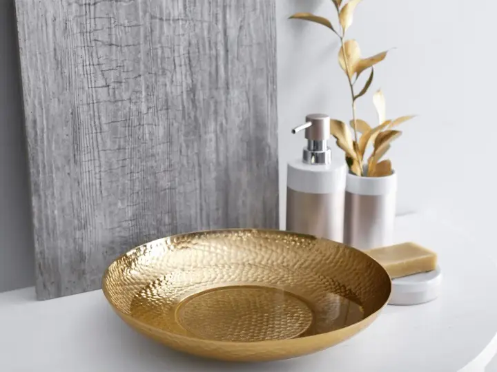 Un bol martelé doré, un distributeur de savon et une barre de savon sur un comptoir blanc, avec une plante dorée et un panneau texturé gris en arrière-plan. Une touche de luxe pour votre douche dorée.