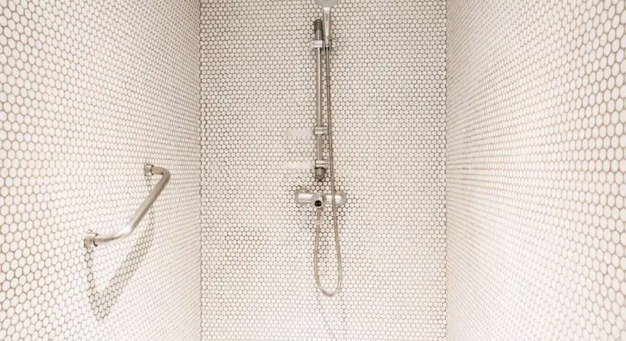 Douche moderne avec carrelage blanc en mosaïque. Une barre d'appui chromée et un pommeau de douche argenté se détachent sur le fond. Le receveur noir est discret.