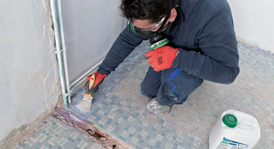 Préparation d'un ragréage sur carrelage ancien. Un ouvrier, masqué et ganté, applique un primaire d'accrochage au pinceau sur le sol carrelé.