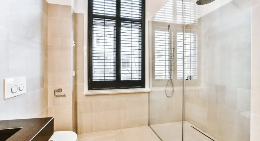 Salle de bain moderne avec douche à l'italienne, carrelage beige, fenêtre à persiennes noires et WC suspendu.
