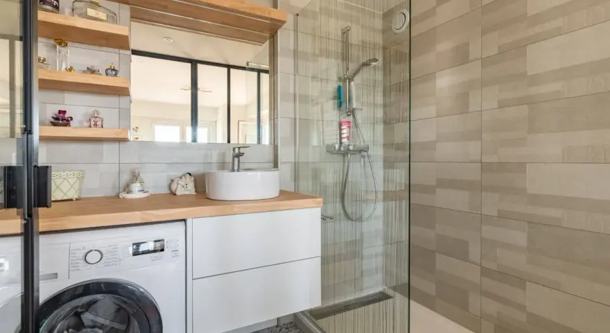 Coin buanderie dans salle de bain moderne avec lave-linge, lavabo, douche vitrée et étagères décoratives.