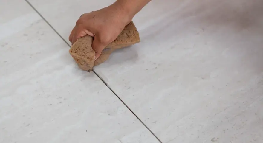 Main tenant, quelle couleur de joint pour carrelage beige ? Une main utilise une éponge pour nettoyer les joints entre des carreaux beiges clairs.