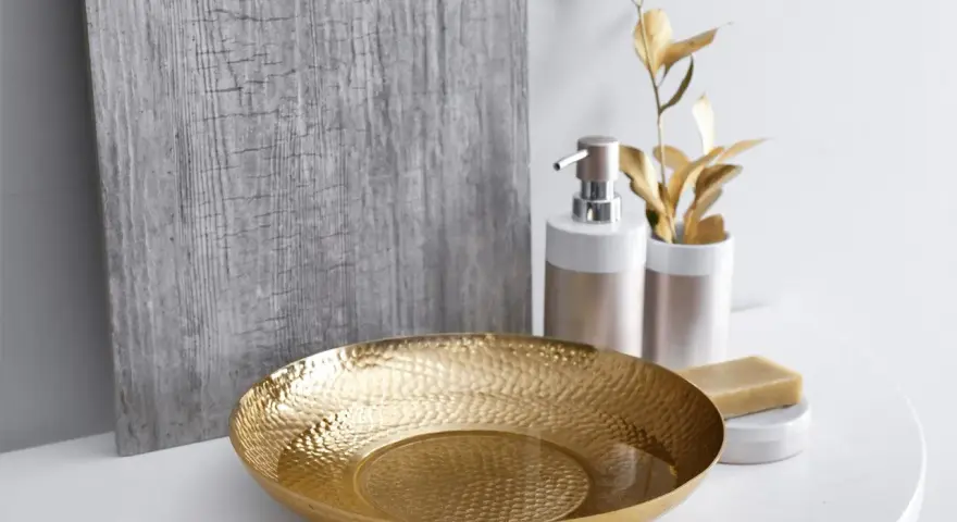 Un bol martelé doré, un distributeur de savon et une barre de savon sur un comptoir blanc, avec une plante dorée et un panneau texturé gris en arrière-plan. Une touche de luxe pour votre douche dorée.