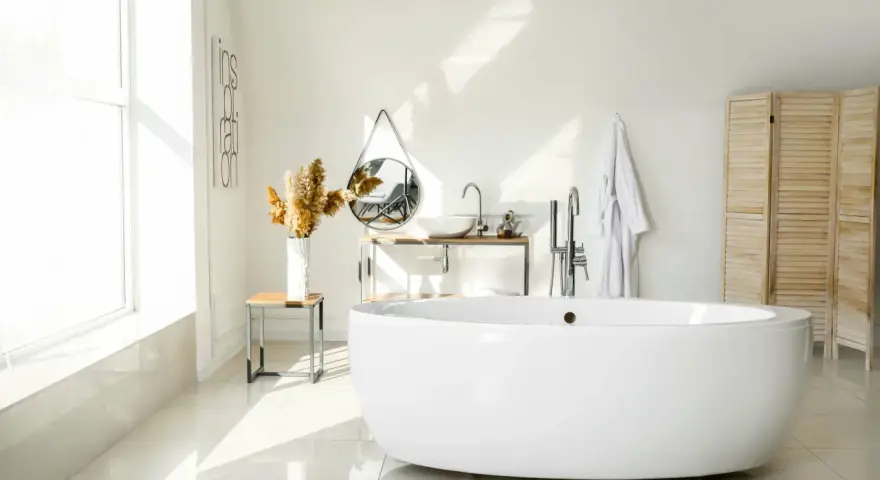 Salle de bain lumineuse avec baignoire îlot blanche, lavabo, miroir, porte-serviettes et paravent en bois. La largeur d'une baignoire est parfaite pour ce coin détente.