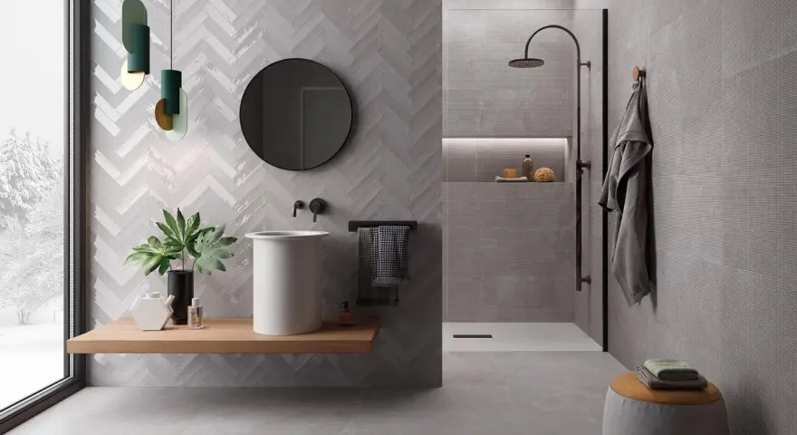 Salle de bain moderne avec murs en **grès cérame** à chevrons et texturés. Lavabo suspendu, douche à l'italienne, miroir rond et touches végétales.