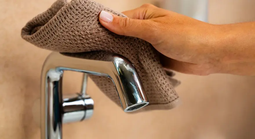 Main d'une femme nettoyant un mitigeur de douche avec un tissu marron.
