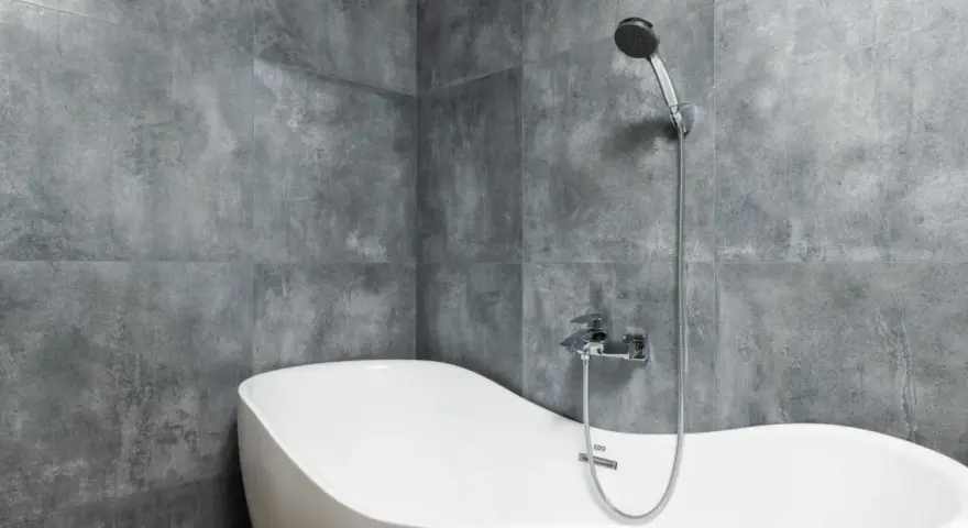 Baignoire blanche aux formes douces, adossée à un mur de carrelage gris en pose en quinconce. Douchette chromée au-dessus.