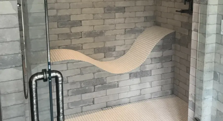 Douche moderne avec un banc ondulé intégré dans le mur de briques. La poignée de la porte en verre est en métal vieilli.