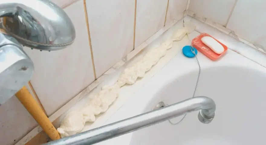 Mousse expansive appliquée pour nettoyer joint plastique porte douche. Savon et bouchon de bain à proximité.