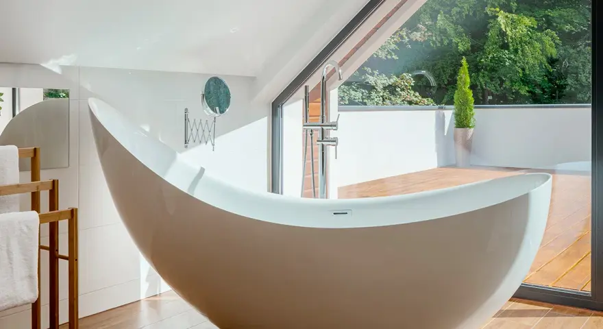 Baignoire design en matière composite, avec robinetterie chromée, devant une grande baie vitrée donnant sur un jardin luxuriant.