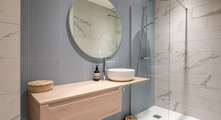 Aménagement petite salle de bain : meuble suspendu en bois clair, vasque ronde, miroir rond, douche à l'italienne.