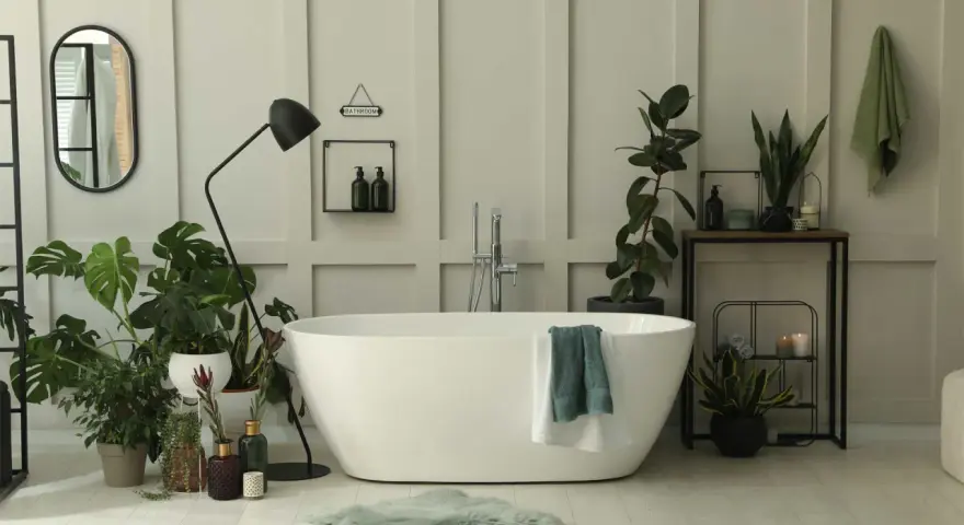 Une salle de bain baignée de lumière, avec une baignoire blanche, de nombreuses plantes vertes qui prospèrent dans l'humidité, et un miroir ovale.