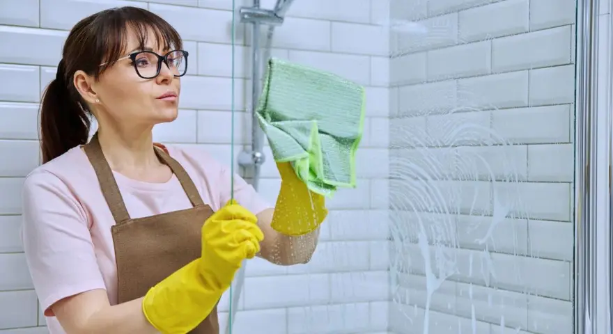Femme avec des gants jaunes et une lingette verte en train de nettoyer le calcaire de la douche.
