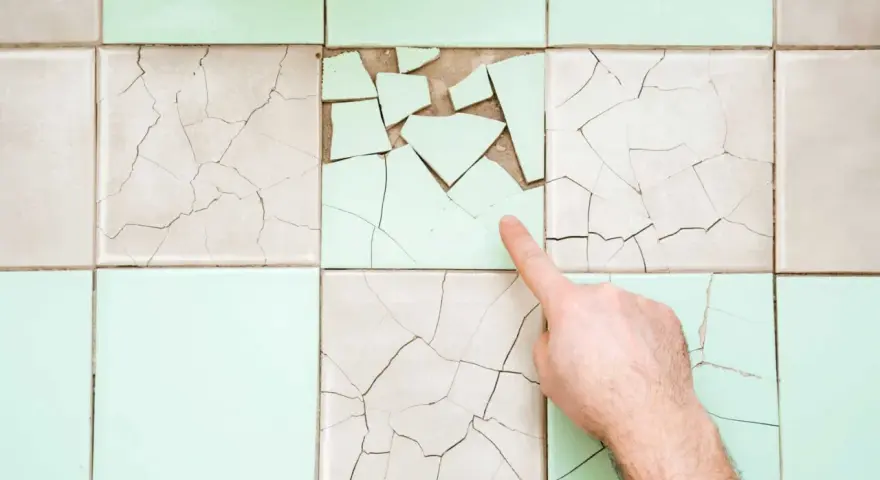 Un doigt pointe une **fissure carrelage** sur un mur de carreaux anciens, certains brisés, d'autres craquelés.