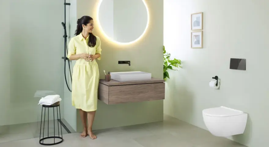 Une femme en robe jaune admire une salle de bain moderne. Le mur vert pâle est une tendance couleur mur toilette, complété par un meuble vasque en bois et un WC suspendu.