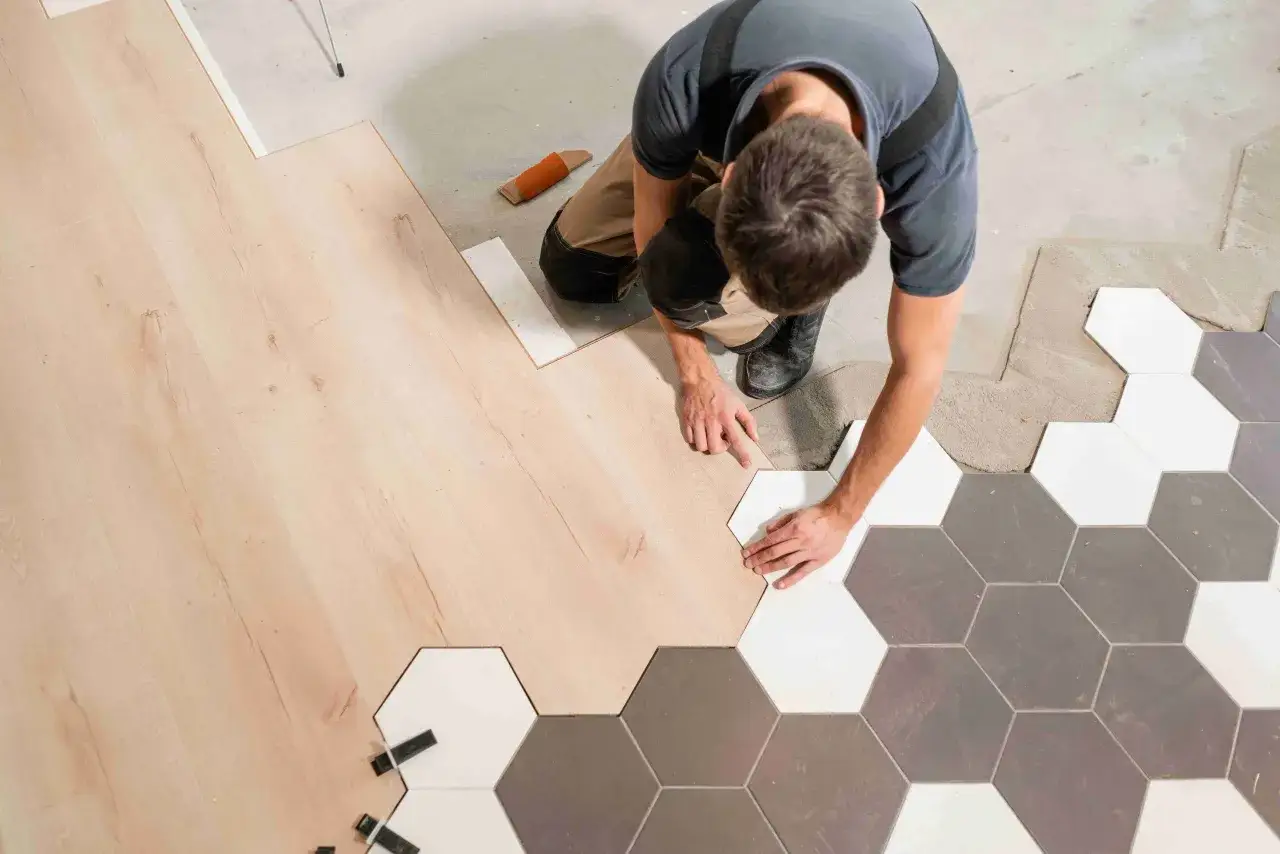 Un ouvrier pose du carrelage hexagonal blanc et gris à côté d'un parquet en bois clair.