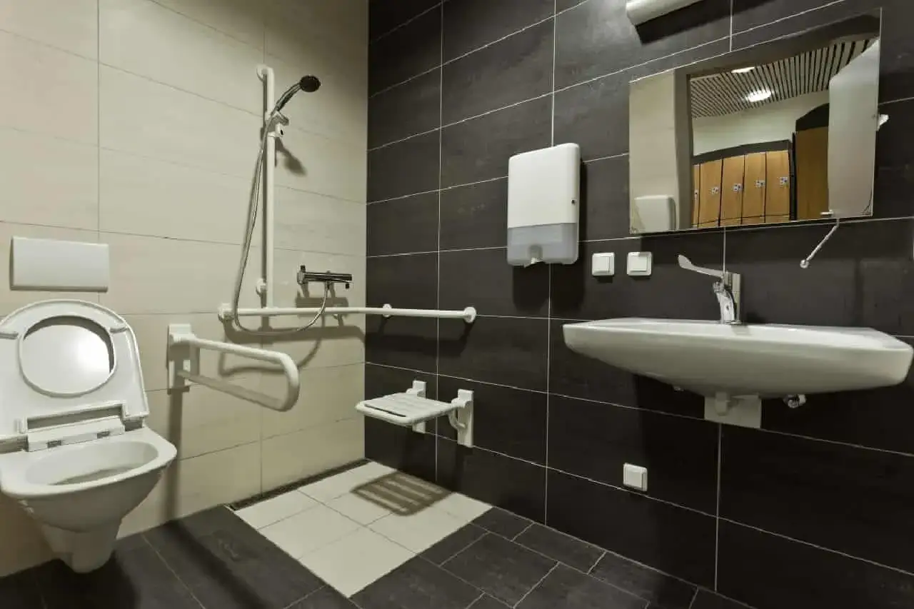 Salle de bain PMR avec cuvette, douche, lavabo, barres d'appui et siège rabattable. Confort et sécurité assurés.