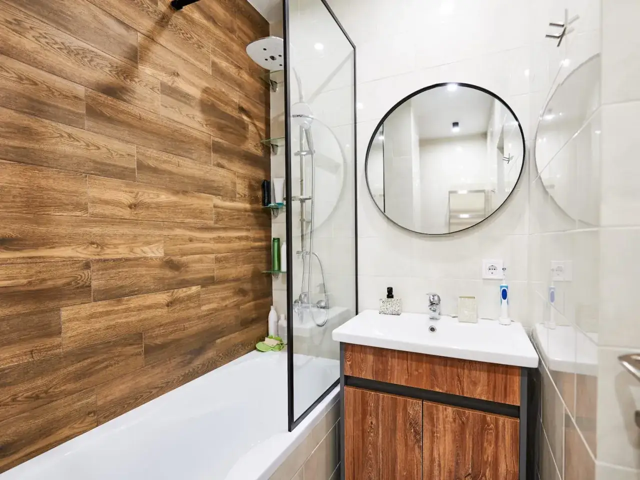 Aménagement petite salle de bain : baignoire avec mur imitation bois, paroi de douche en verre, lavabo sur meuble bois, miroir rond.