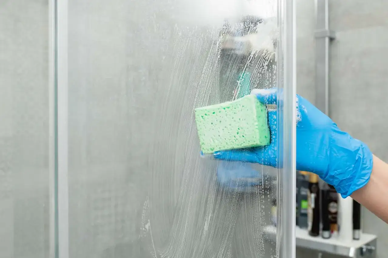 Main tenant, une main gantée de bleu nettoie une paroi de douche en plexiglas avec une éponge verte savonneuse.