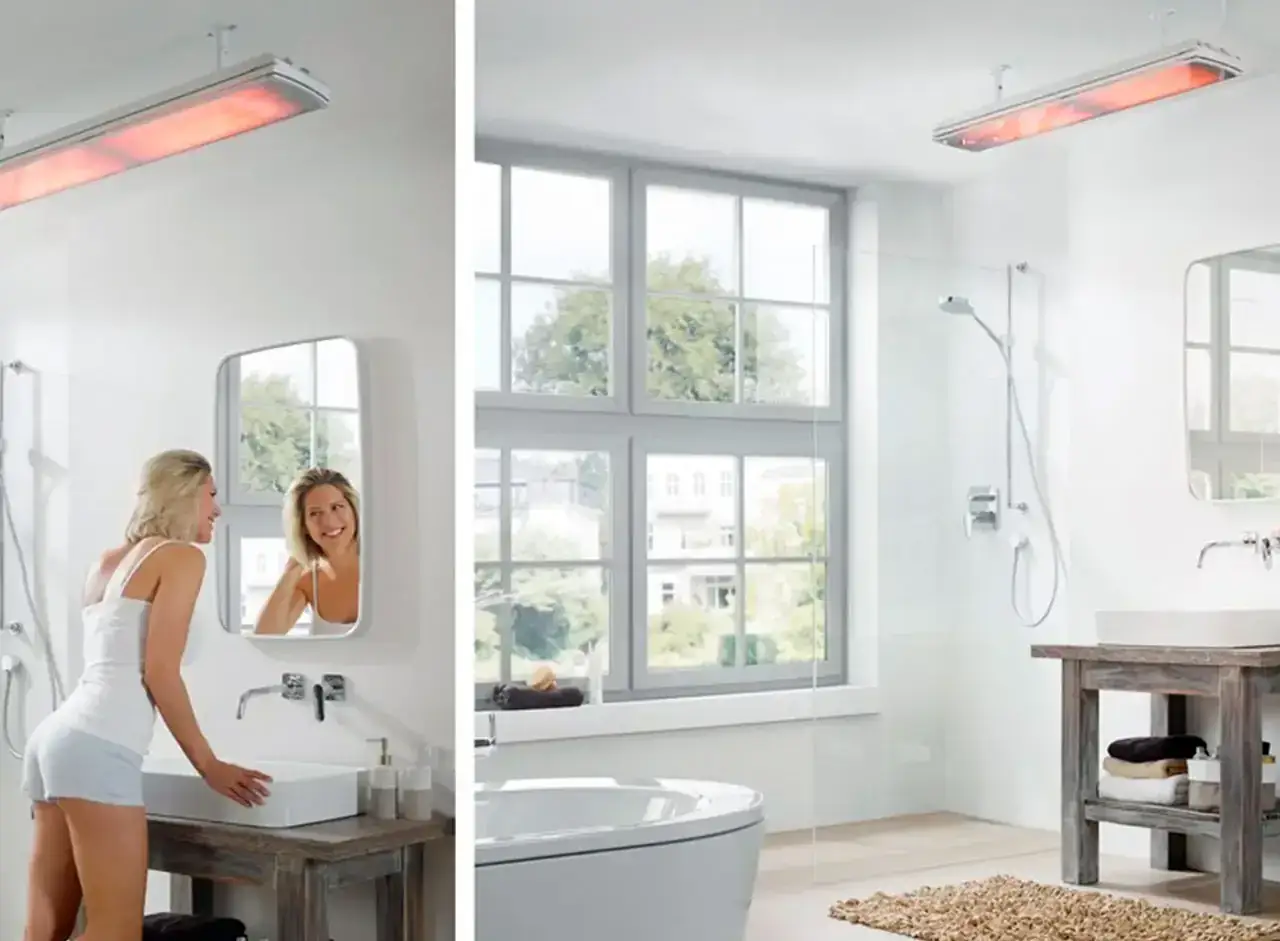 Une femme souriante se regarde dans le miroir d'une salle de bain moderne. Au-dessus, un appareil de chauffage infrarouge, une chaudière ou chauffe eau, diffuse une douce chaleur rouge.