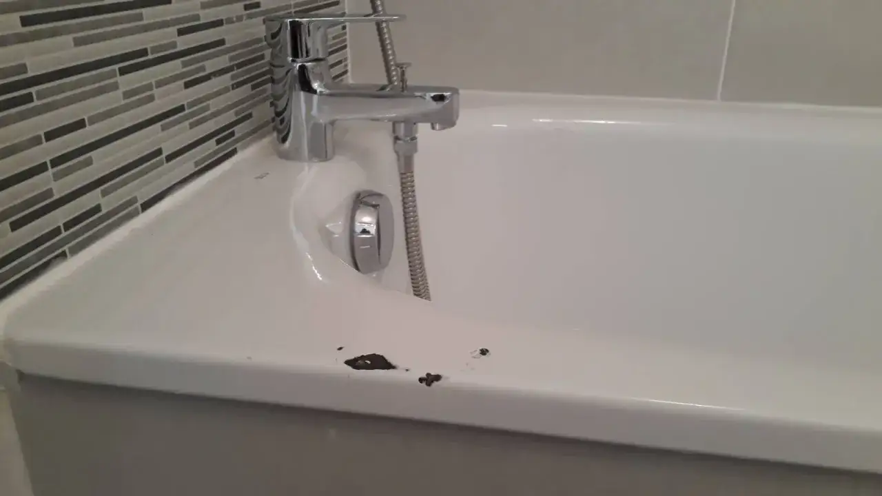 Bain en émail blanc avec une rayure sombre et des éclats sur le bord. Robinet chromé et carrelage mural à rayures grises en arrière-plan.