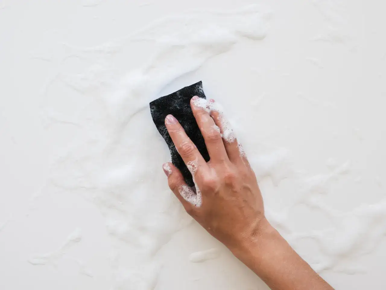 Main tenant, une main frotte avec une &eacute;ponge noire sur une surface blanche savonneuse, illustrant comment nettoyer un bac de douche en r&eacute;sine.
